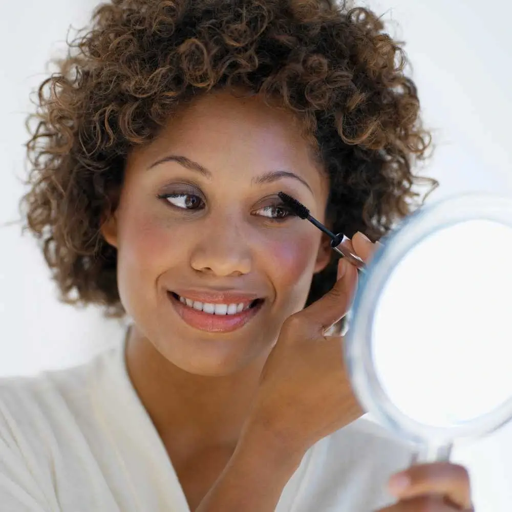 woman with curly hair applying mascara while holding a mirror