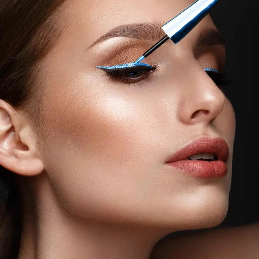 closeup portrait of a woman with makeup applying blue glitter eyeliner on her eyelid