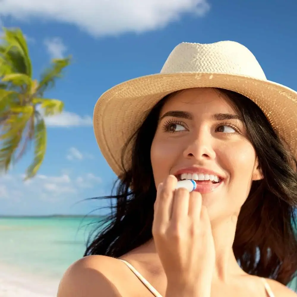 closeup portrait of a woman wearing a beach hat applying lip balm on the beach