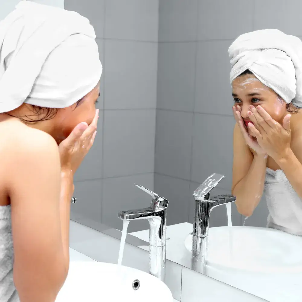 young woman with a towel wrapped on her head washes face in front of the mirror