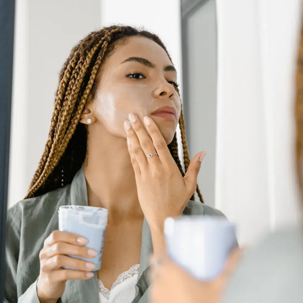 young woman with braided hair applying face moisturizer in front of the mirror
