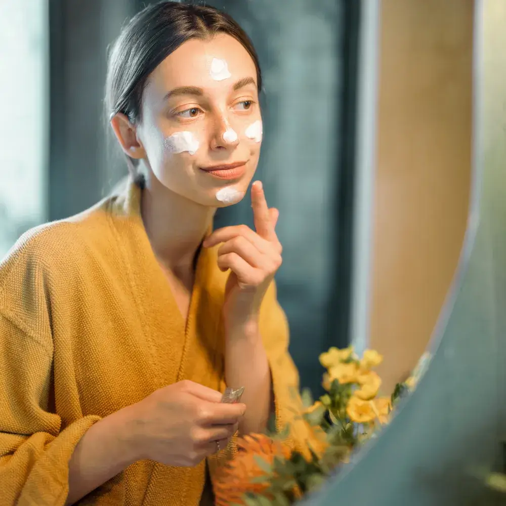 woman wearing a bathrobe in front of a mirror applying face cream