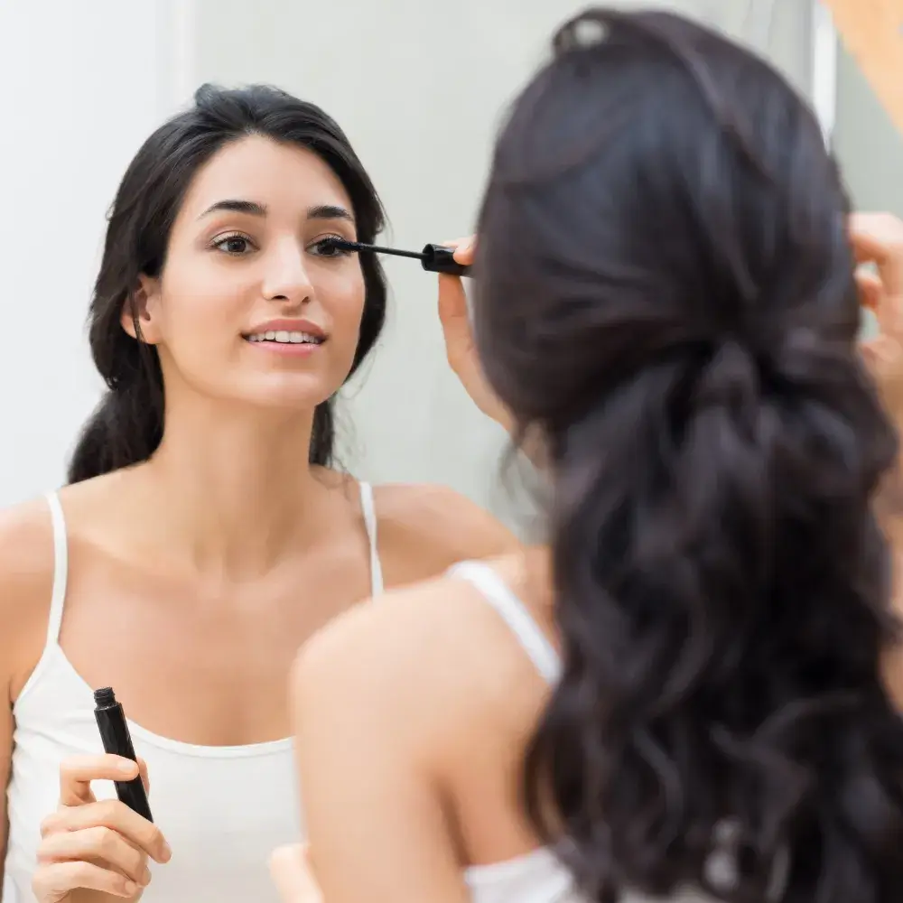 young woman wearing a white top applying mascara in front of a mirror
