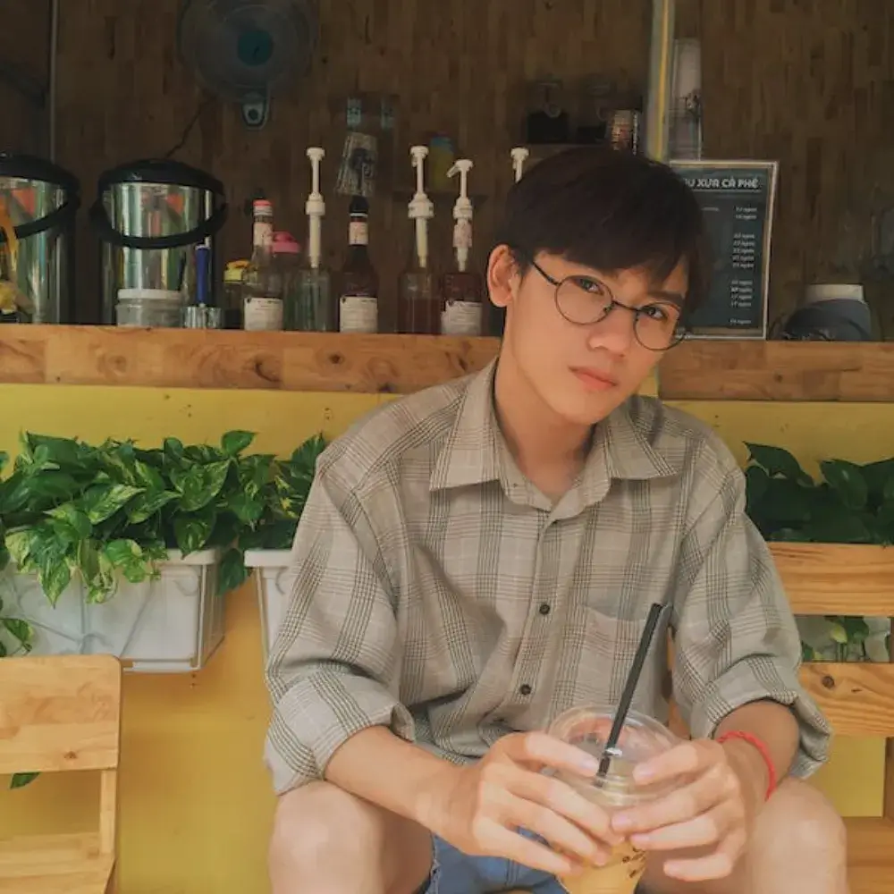 asian young man wearing glasses sitting in front of a food stall holding a drink