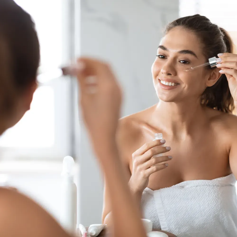 Young woman in a white towel applying face oil while standing in front of a mirror