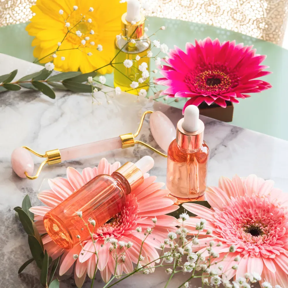 Two face oil bottles and a rose quartz roller on a marble table, surrounded by vibrant flowers