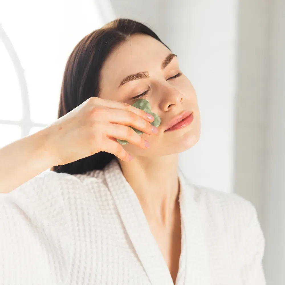 Young woman in a white robe using a gua sha to massage her face