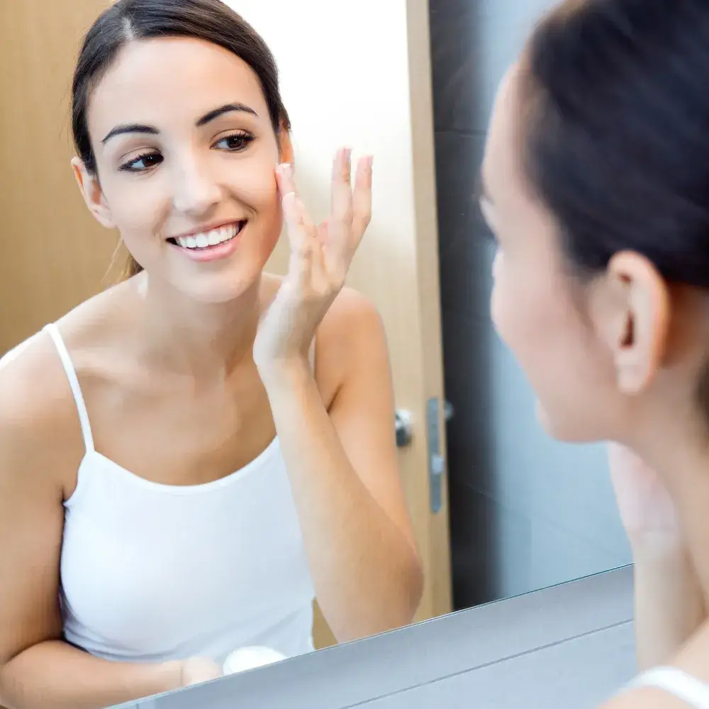 young woman applying a moisturizer on her face in front of a mirror
