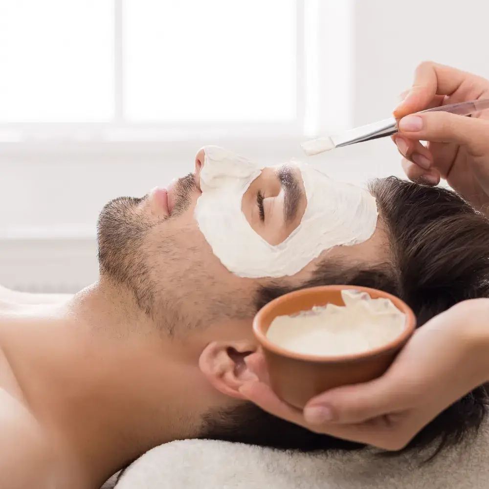 A man with facial hair receiving a facial mask treatment from a beautician at a salon