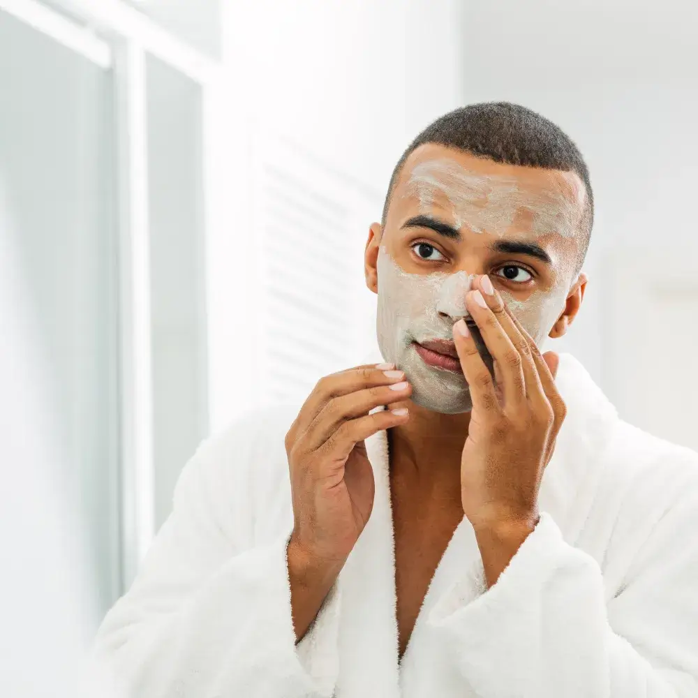 Young man applying a clay mud mask to his face in front of a mirror