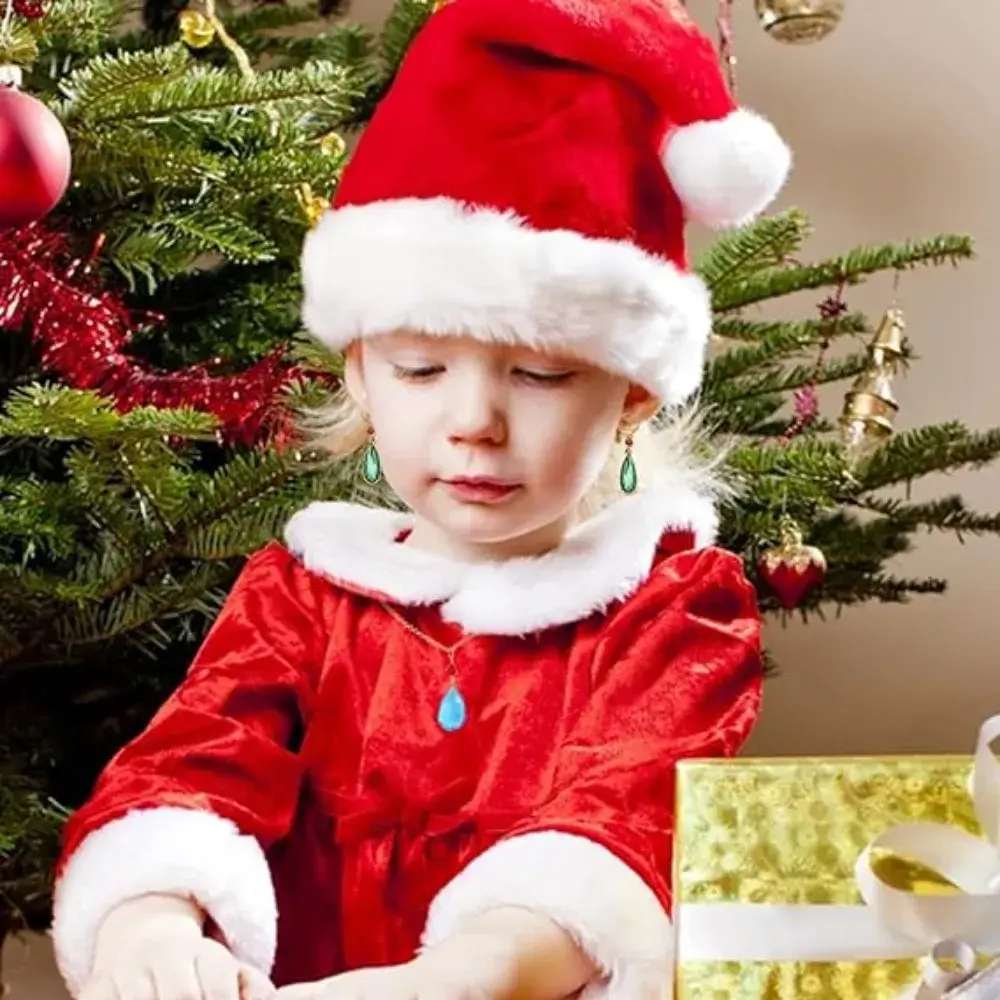 A little girl dressed in a Santa Claus costume wears a sky-blue howl's necklace, with a Christmas tree behind her.