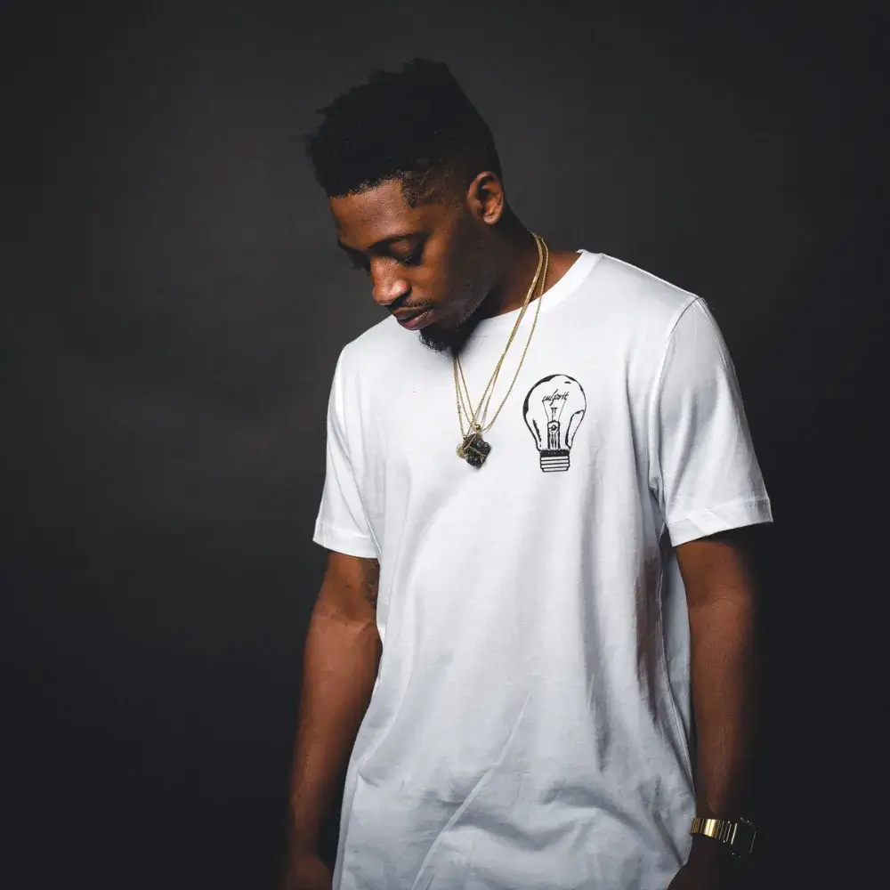 young man in a white shirt wearing a watch and gold necklaces against a black bakground