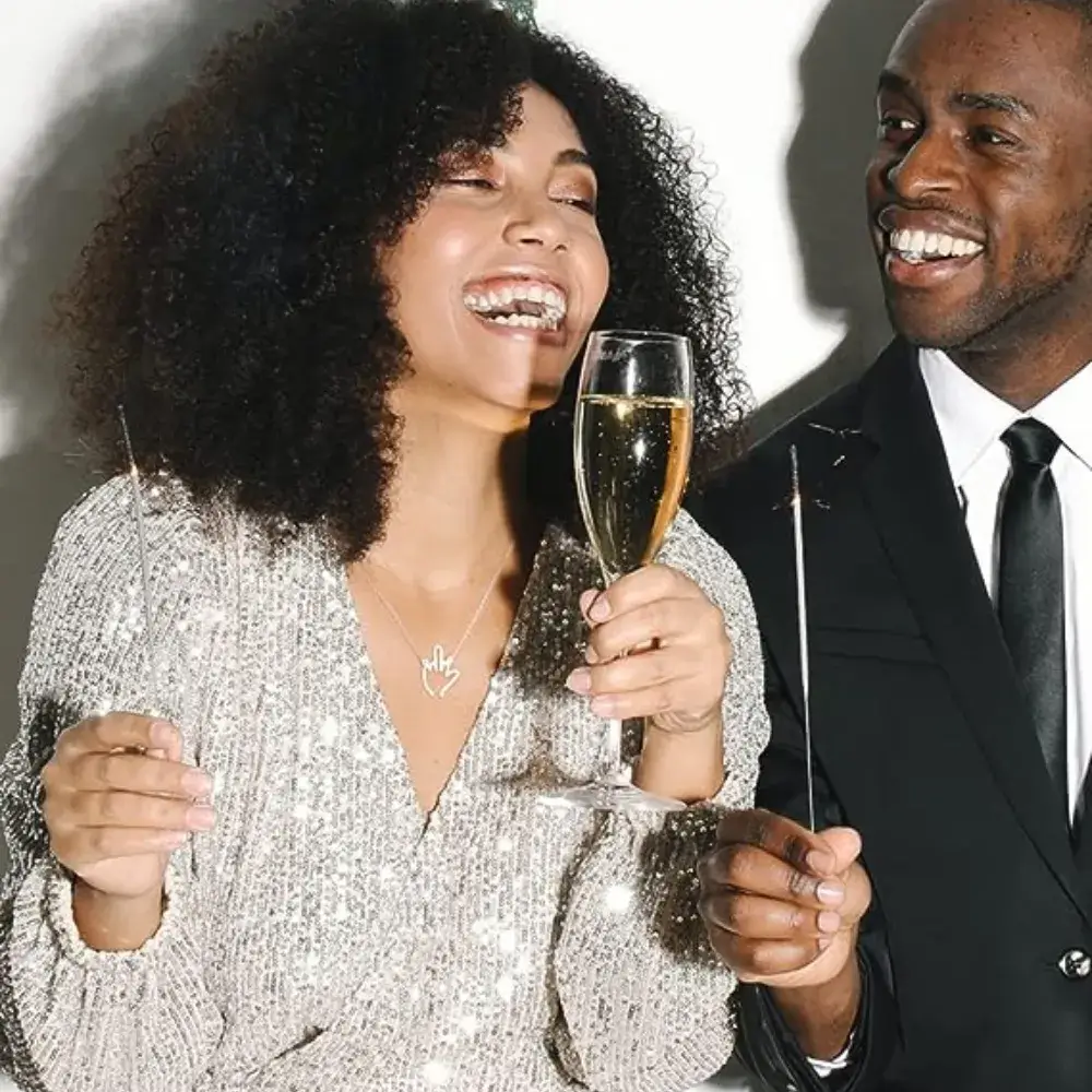 A woman in a silver dress, adorned with a necklace featuring a silver middle finger pendant, is laughing alongside a man.