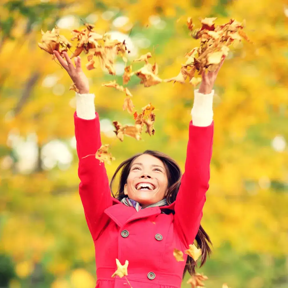 happpy woman wearing a red coat throwing leaves up in the air with arms raised