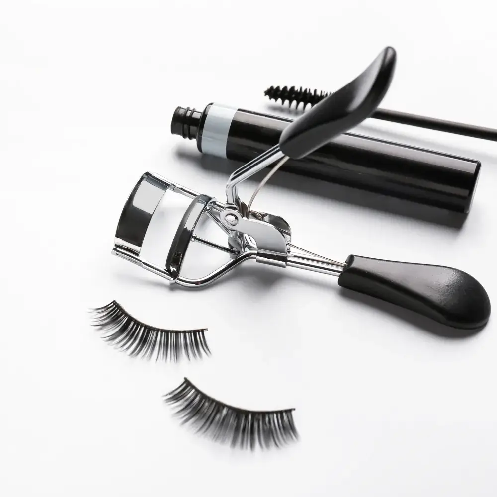 black mascara, eyelash curler and a pair of false eyelashes against a white background