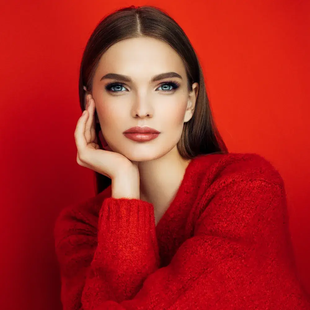 A beautiful brunette woman with makeup, wearing a red sweater, posed against a red background