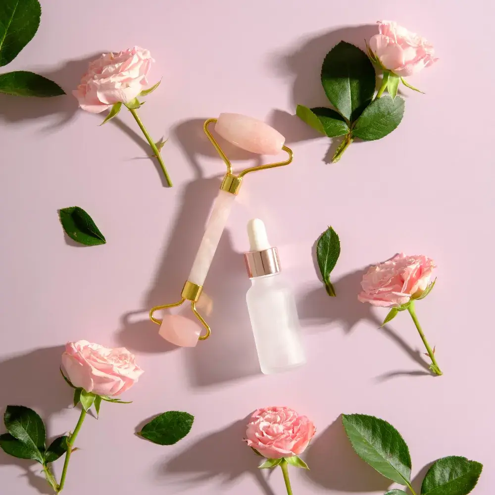 face oil and rose quartz face roller surrounded  by rose buds on a pink backround