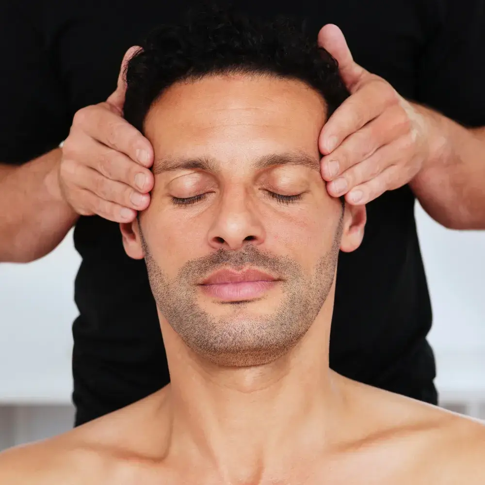 portrait of a man with his eyes closed getting a face massage from a masseur