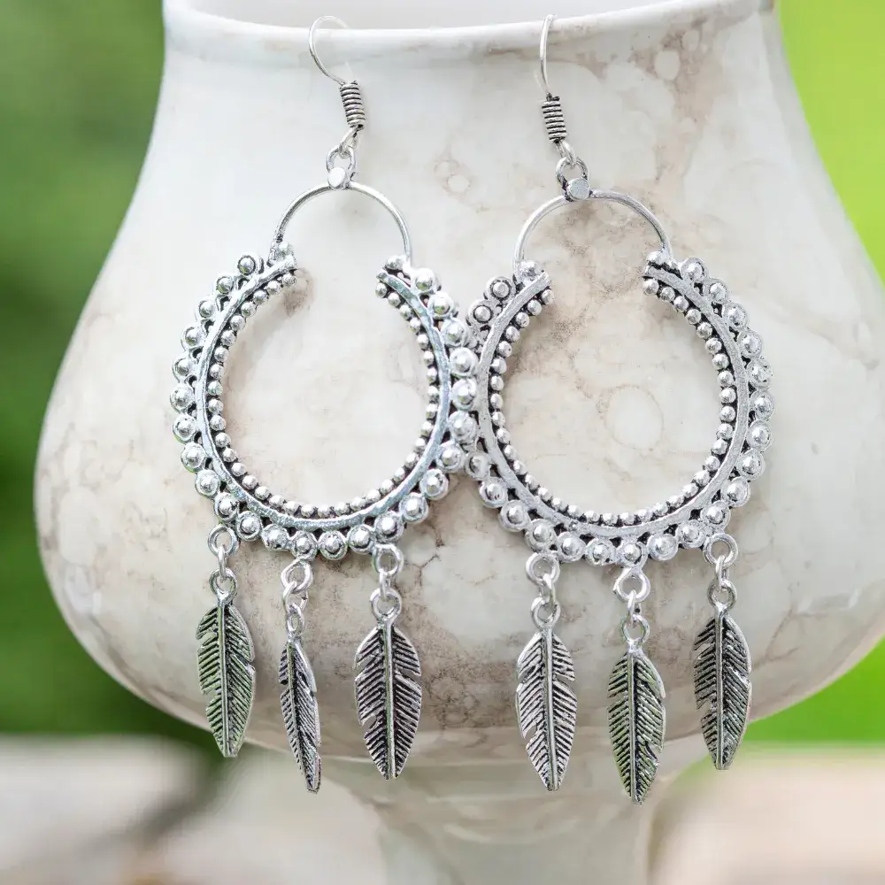 Silver earrings with a dream catcher design are hung on a cup against a green background
